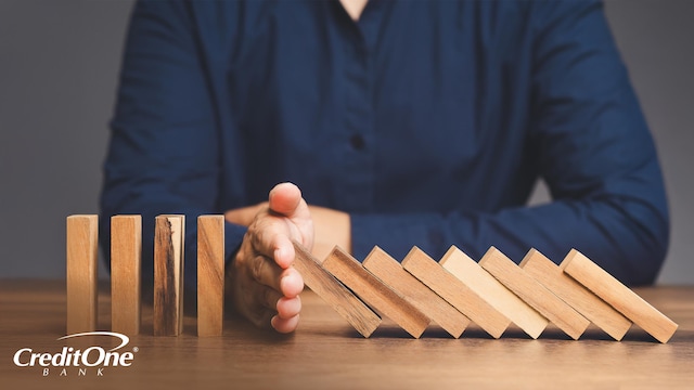 A man uses his hand to stop wooden blocks from toppling over like dominos, representing the impact of poor debt management on credit scores and taking action to prevent it.
