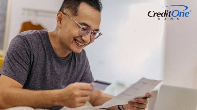 A man holds a credit card in one hand as he reviews a document he’s holding in the other. He is smiling, perhaps because this is the first step he’s taking toward rebuilding his  credit with credit cards.