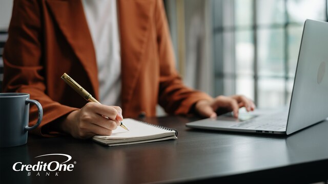 A woman, framed from the neck down, sits at a desk with coffee and a laptop while writing in a spiral notebook. She looks as if she may be checking items off a list, a useful tool for staying on top of credit card payments.