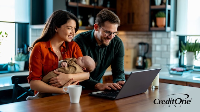 A couple reviews their finances on a laptop at the kitchen table, with the mother holding their newborn. They may be working on the family’s financial planning.